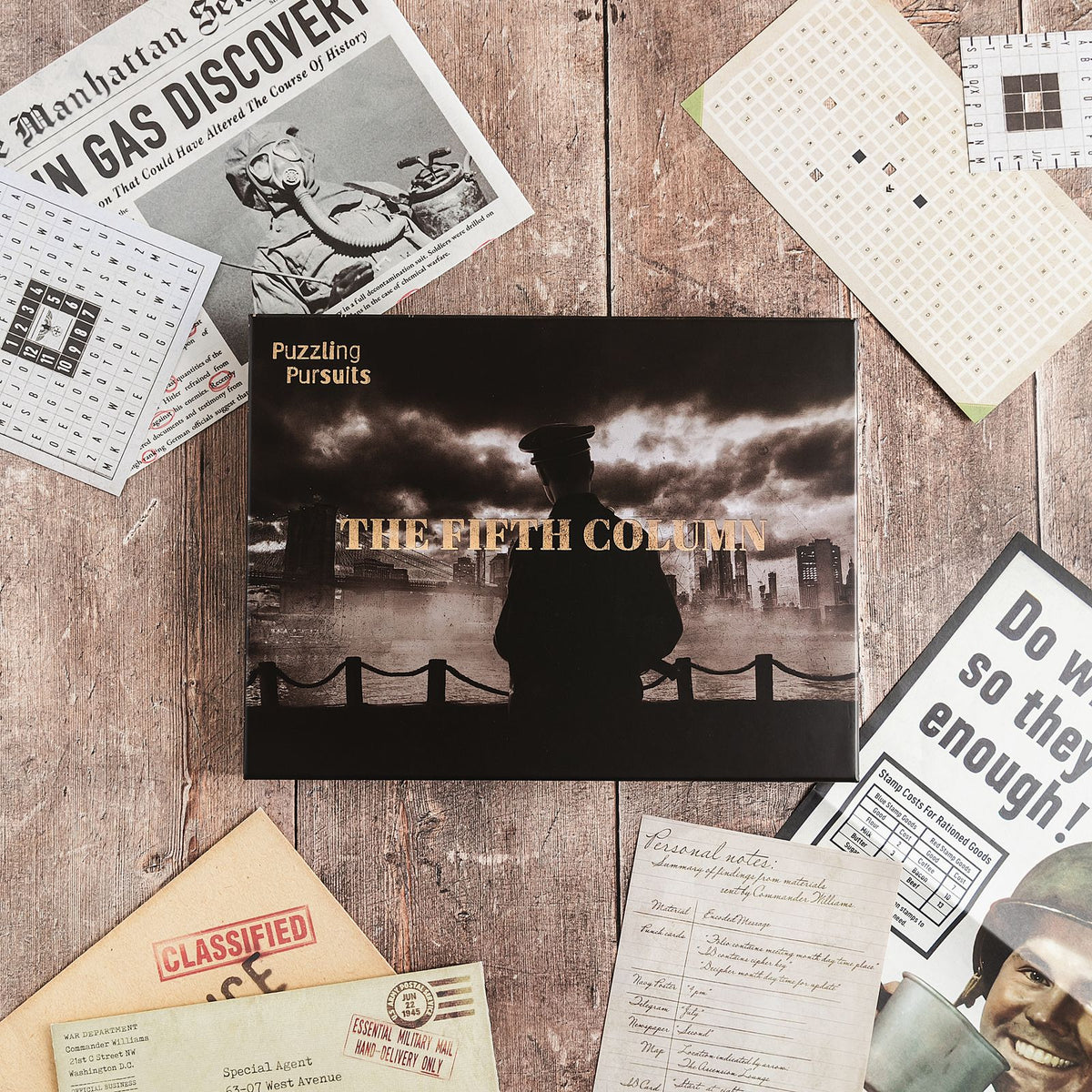 The Fifth Column game box on a wood table surrounded by a Manhattan newspaper page, punched decoder cards, a wartime poster and a kraft envelope stamped CLASSIFIED