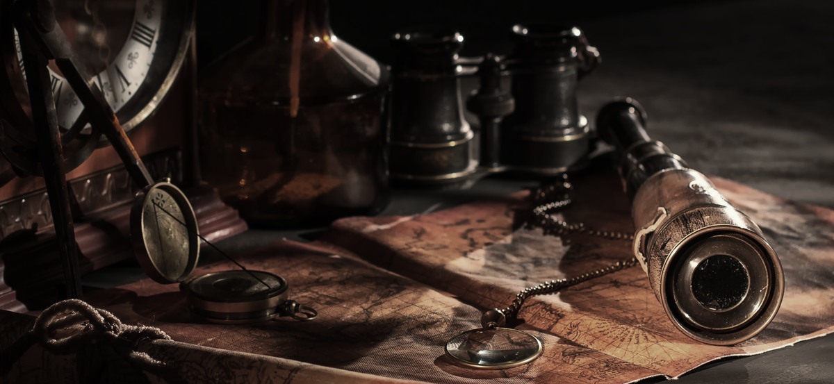 An antique desk lit from one side: a clock face, a glass apothecary bottle, brass binoculars, a spyglass resting on an old map, a compass and a magnifying lens on a chain