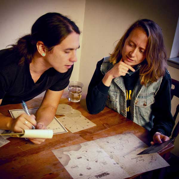 Two players at a wooden table working through Blackbrim: 1876, one leaning in to write notes with a pen while the other rests their chin on their hand and studies the materials, with the Town of Blackbrim map and a cipher wheel card spread out in front of them